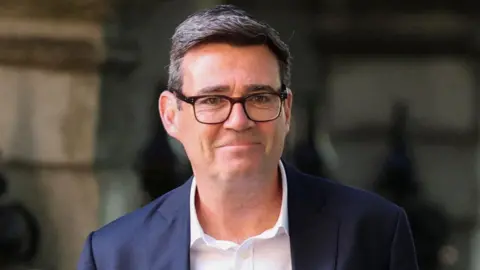 Portrait of a person wearing dark-framed glasses, a navy blazer, and a white shirt, standing outdoors in front of a blurred stone building background. The person is facing the camera with a neutral expression.