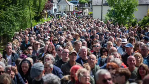 Thousands of people walking down a village street with houses in the background and green bushes on either side.