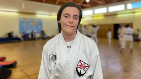 BBC Grace Greenwood smiles at the camera in front of karate class while wearing her Jersey Wado Ryu gi