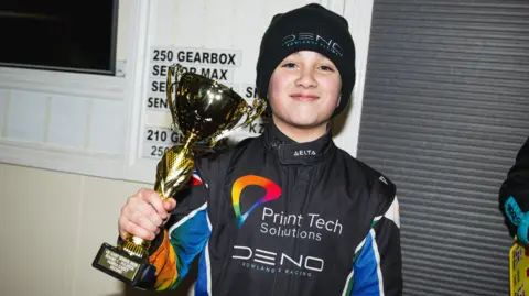 Josh East Photography Deno wearing a black beanie hat with his name on it, and black racing overalls with his sponsor on the chest. He is smiling at the camera and holding up a gold trophy in his hand.