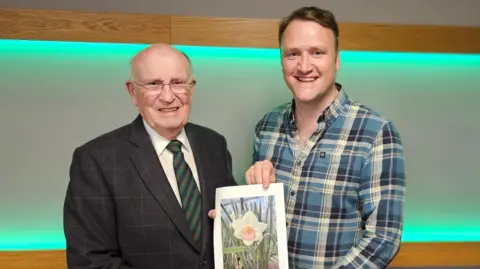 David Maxwell An elderly man in a dark suit and green striped tie stands to the left of another man in a chequered blue shirt. They are both holding a photograph of a daffodil.