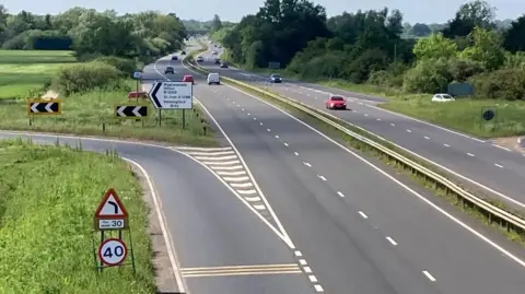 A dual carriageway with a slip road off to the left. A 40mph sign is visible at the entrance to the slip road, with a left-hand bend sign above. There is also a signpost to Biggleswade, St Ives and other places as the left-hand bend begins. There are grass and trees on either side of the road, and some traffic is visible on both sides.