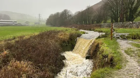 Ned/BBC Weather Watchers A river in Ulverston, Cumbria. Drizzle comes down on to fields and a river is flowing in the centre of a ditch. 