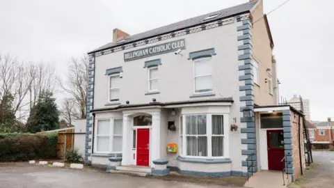 CAMRA The exterior of the Billingham Catholic Club. The facade is mostly white with some blue accents. It has a bright red door in the middle and a brown door on the right hand side. The building is two storeys.