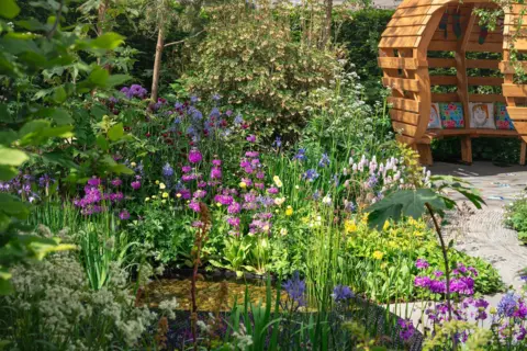 Julie Skelton A close up of the pond which represents the barriers people with Down's syndrome face in society. Tall plants with purple and pink flowers surround the path.