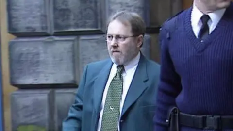 Andrew Walker outside court with a prison guard. He is wearing a green suit and tie and glasses.