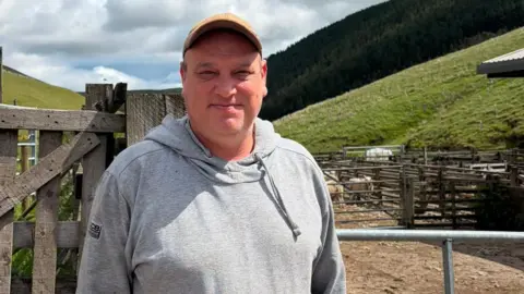 BBC/Grace Stewart-Piercy Matt Ridley, a middle-aged man wearing a grey hoodie and a yellow baseball cap, stood on his farm. In the background is a steel gate, leading to separate sheep pens. Steep, green hills rise upwards in the background.