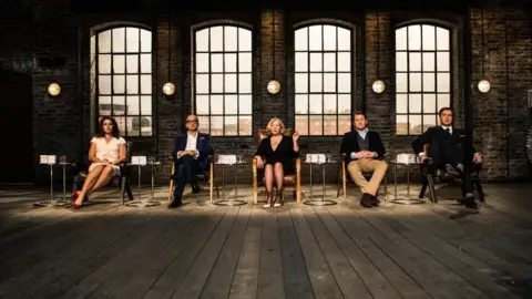 A row of judges from Dragon's Den sit in a row in separate chairs in a warehouse. There is a spotlight on all five. 