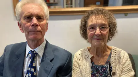 A man and a women sitting next to each other and looking at the camera. The woman is wearing a flowery blouse, a white cardigan and a pair of glasses, whilst the man is wearing a blue blazer and tie and a white shirt.