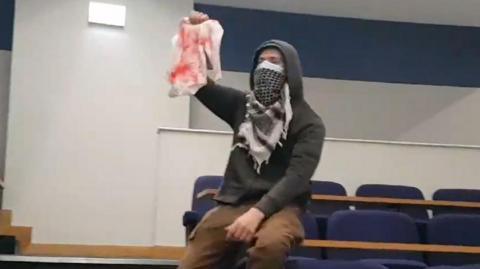 A protester sitting on a lecture hall desk while holding baby clothes covered in fake blood