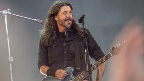 Getty Images O músico norte-americano Dave Grohl, da banda de rock Foo Fighters, se apresenta no Orange Stage do Roskilde Festival em Roskilde, Dinamarca, em 5 de julho. Grohl veste uma camisa preta e está com a mão direita no topo do violão enquanto posa antes da apresentação. Grohl olha para a multidão.