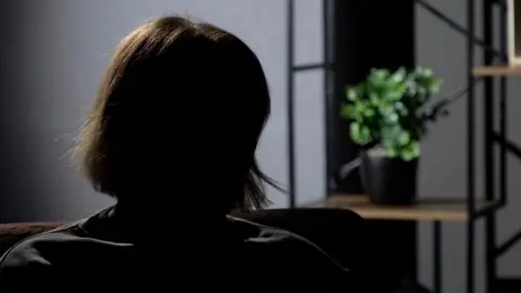 A woman sits in silhouette to protect the interviewee's anonymity. She is sitting in front of a grey background with plastic flowers on a wooden shelf. 