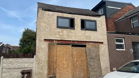 A boarded up house with a flimsy roof and two old windows