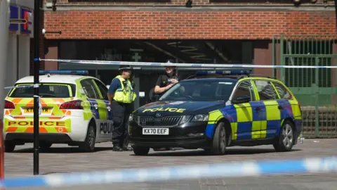 Two police cars are parked behind a blue and white police cordon. Two police officers stand between the cars talking to each other.