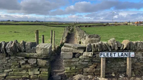 A road sign spelling out Fleet Lane in front of dry stone walls and a footpath between green fields 