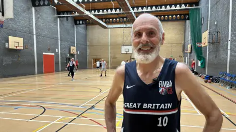 Gordon Shovlin wears a navy blue vest with Great Britain written on the front in white lettering. He is bald with a white beard. He is pictured standing in a sports hall while other men play basketball behind him.