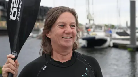 Sarah Perkins is holding an ore and wearing a wetsuit. She is standing near the water and smiling.