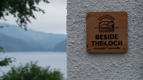 A close up of a wooden sign saying "Beside the loch", the couple's business. To the left of the wall where it is hung, we see beyond the building to the spectacular loch view, framed by trees.