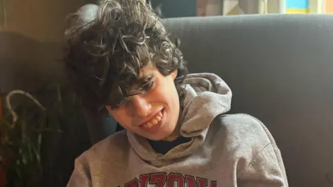 Family handout A picture of a boy, looking down and smiling. He has brown curly hair and is wearing a grey hoodie.