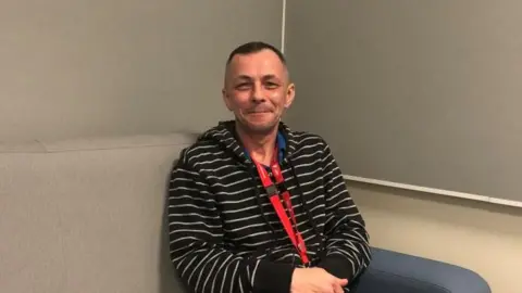 Vikki Irwin/BBC Dr Granville Sutton sits on a sofa in an interview room in at the Martlesham Police investigation centre. He's sitting on a comfortable sofa wearing a black and white stripped jumper and dark trousers.  