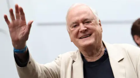 Actor John Cleese is seen smiling and waving at the camera. He has white hair and is wearning a cream suit jacket, blue watch and black jumper.