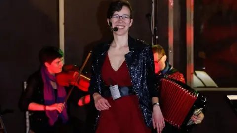 Michael Heath A person with short dark hair wearing a headset microphone and smiling at the camera wearing a red dress and dark shawl. There is a person behind her playing an accordion and another playing a violin.