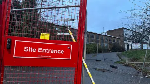 BBC A red gate on the left hand side with a sign saying Site Entrance and more metal fencing in front of the derelict police station site with boarded up windows.