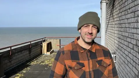 Nik Mitchell standing on the roof of Arlington House in Margate next to a wooden nesting box which overlooks the sea