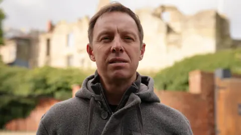 The head and shoulders of a man with short brown hair and a grey coat. He is standing in front of a wall which is out of focus, behind the wall is a hedge, and further back the remains of a castle