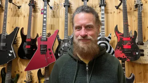 Man with grey hair and a long beard stands in front of a wall of guitars.