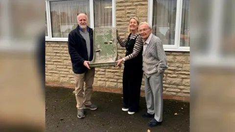 A woman and a man holding up a rusty green pilot seat outside a house, with an elderly man standing next to the woman.