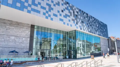The Box A large glass and grey stone building sits in front of a paved area with blue umbrellas. People are sitting outside.