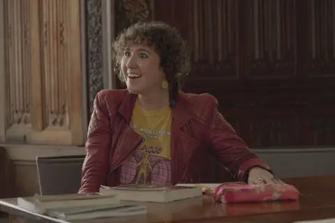 Netflix Esme wearing a red leather jacket with her curly hair in an up-doo, she is wearing a yellow top with a vague logo on and yellow dangling ear rings too match. It's a shot from the movie where she is sitting in class