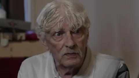 An older man with grey hair and a grey moustache. He is sitting in a room against a white wall. Wearing a white polo shirt.
