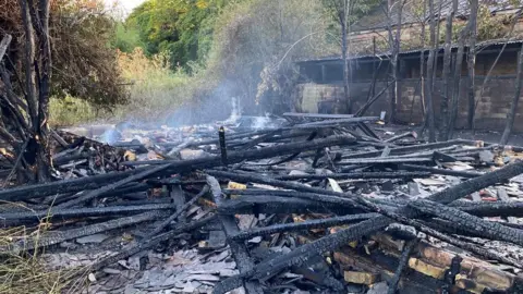 A building which was affected by a fire is now rubble on the floor. There are pieces of wood which have been damaged by the fire, and smoke is rising from part of the rubble to the left. 