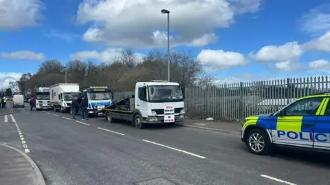 A number of lorries are seen parking on the side of the road in the middle and far ground and closer in the foreground, the side of a Police Service of Northern Ireland jeep can be seen