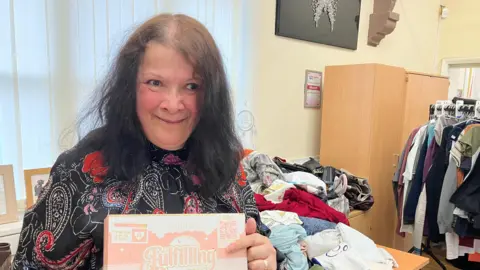 James Grant/BBC A woman in a black dress with red and white patterning stands in front of a table with lots of jumbled up clothing on it. She is holding a box with clothing in it. 