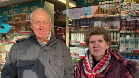 George King/BBC An elderly man with very short white hair wearing a grey jacket standing next to an elderly woman with brown hair wearing a colourful scarf and a purple jacket