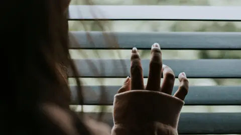 Getty Images Woman looking out the window blinds