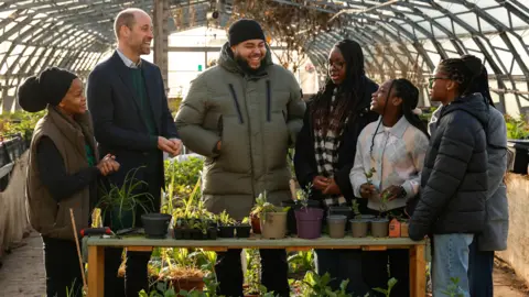 Four teenage girls can be seen stood around a table which has several potted plants on it inside a greenhouse. There is also a man stood in the middle laughing and next to him is Prince William who can be seen looking at the girls and smiling. A woman is stood next to him also smiling.