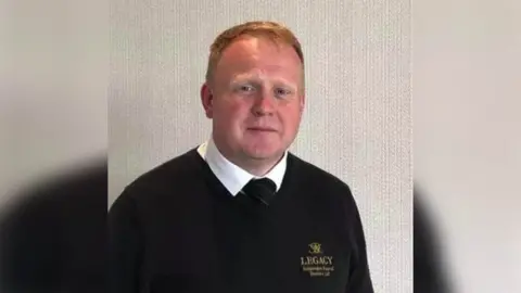 Legacy Independent Funeral Directors Robert Bush has short ginger hair. He is wearing a white shirt, a black tie and a black jumper.