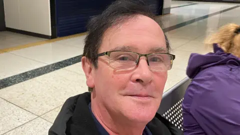 Grace Wood/BBC A man with black hair and rectangular glasses sitting on a metal chair in an empty shopping centre