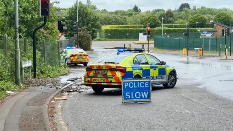 A police car in front of a POLICE SLOW sign. There is water in the road.