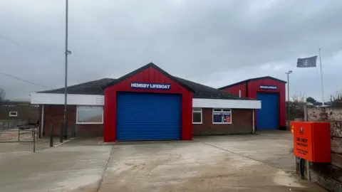 Richard Knights/BBC Hemsby Lifeboat station. It is a brick building with a red front and a large blue sliding door