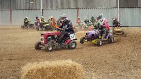 Harry George Hall Lawn mower racers take a corner on the track - there are several competitors in helmets and racing mini tractor-like machines