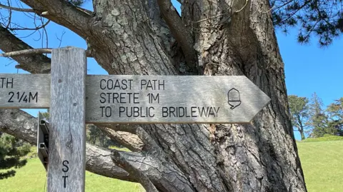 A picture of a wooden sign point pointing the way to the coast path. There is a tree and field behind it .