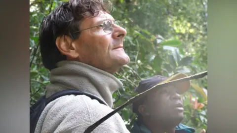 Tom Lovett A man with short curly dark hair and silver-rimmed glasses looks up. He wears a light-coloured fleece. Another man wearing a cap and a blue hoodie stands beside him. Behind him, a dense green forest vegetation can be seen.