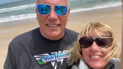 A selfie photograph of Mark and Annette on a beach. Mark is wearing a grey Van Halen t-shirt and clear-framed sunglasses with blue lenses reflecting Annette's hand holding the phone and cars parked nearby. Annette has windswept blonde hair and dark sunglasses. She is wearing a light grey jumper wrapped around her neck. 