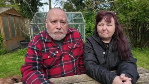 Kevin Morgan and Esta Lippiatt from Camborne sat at a bench in a garden with a shed and greenhouse behind them. Morgan is wearing a red shirt and Lippiatt is wearing a black coat.