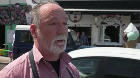 A bearded man in a pink striped shirt and a shop behind.